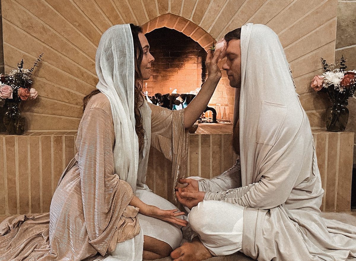 A couple in white robes and veils, one blessing the other